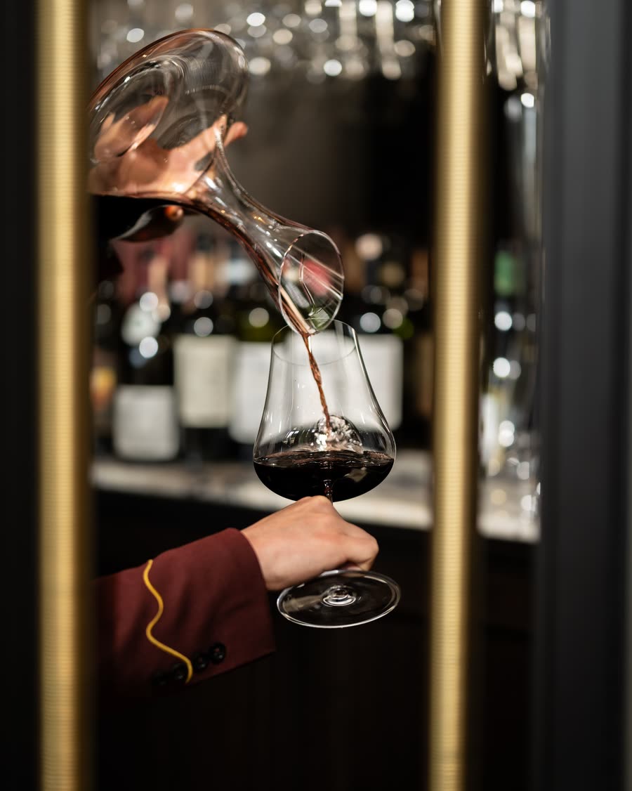 A hand in a dark sleeve pouring red wine into a glass in front of a rack of bottles.