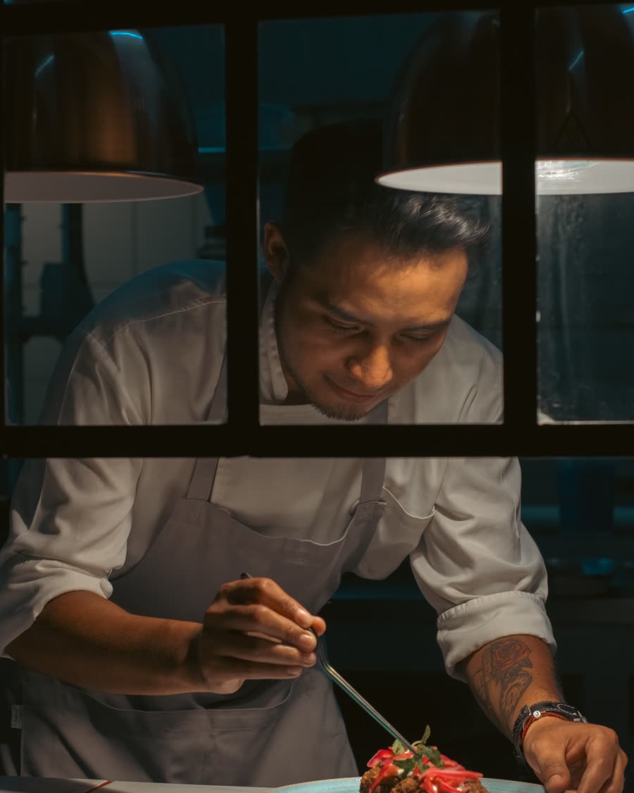 A chef seen through the pass, lit warm from one side, finishing a plate.