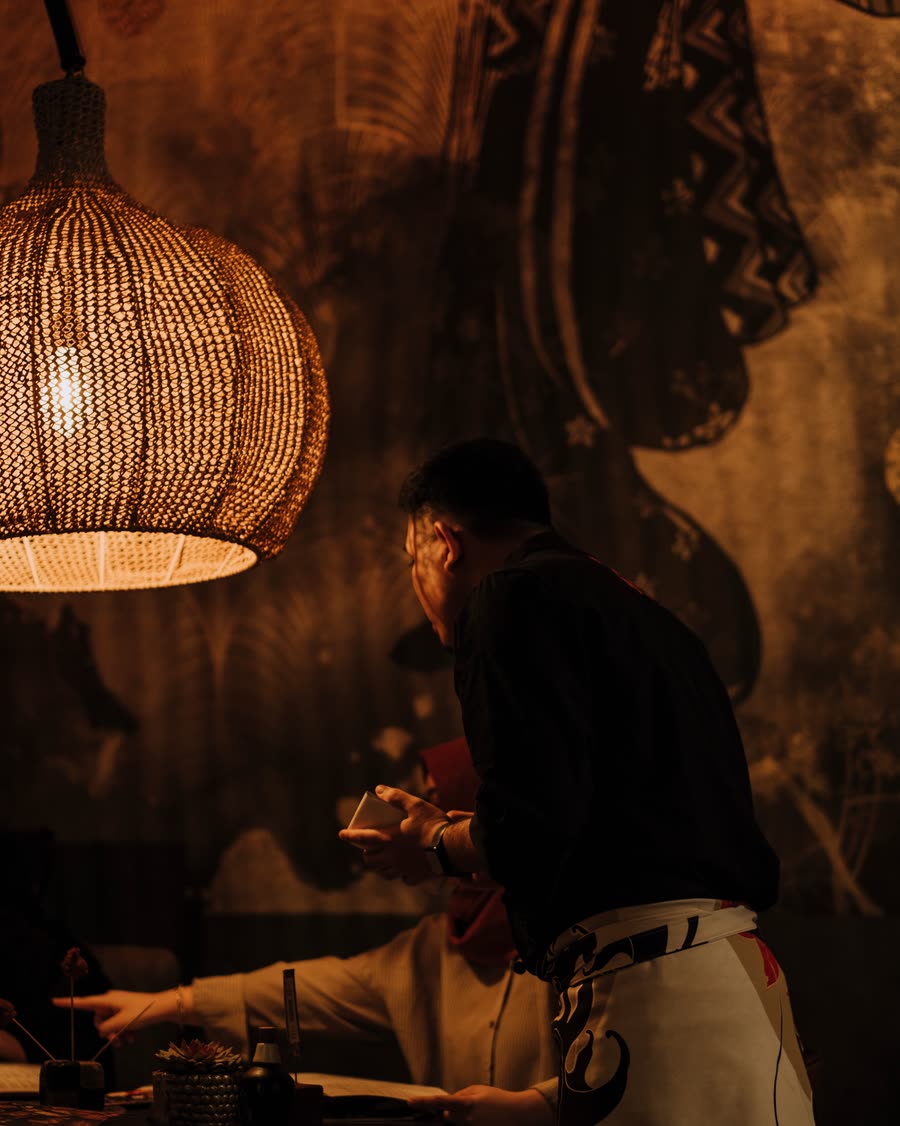 A figure in black serving a table under a woven pendant lamp in a dimly lit room.