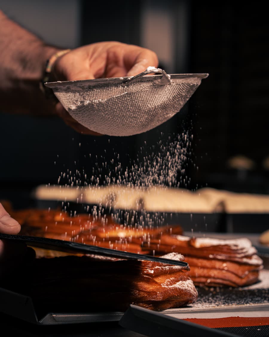 Sugar falling through a sieve over a tray, backlit against a dark kitchen.