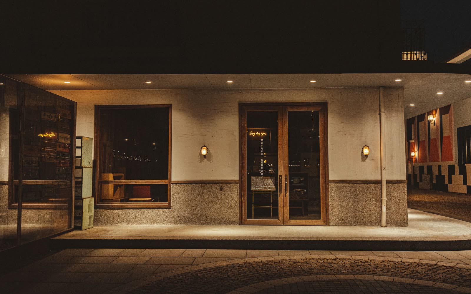 A restaurant frontage at night: two warm windows, wooden doors and wall lamps over wet cobbles.