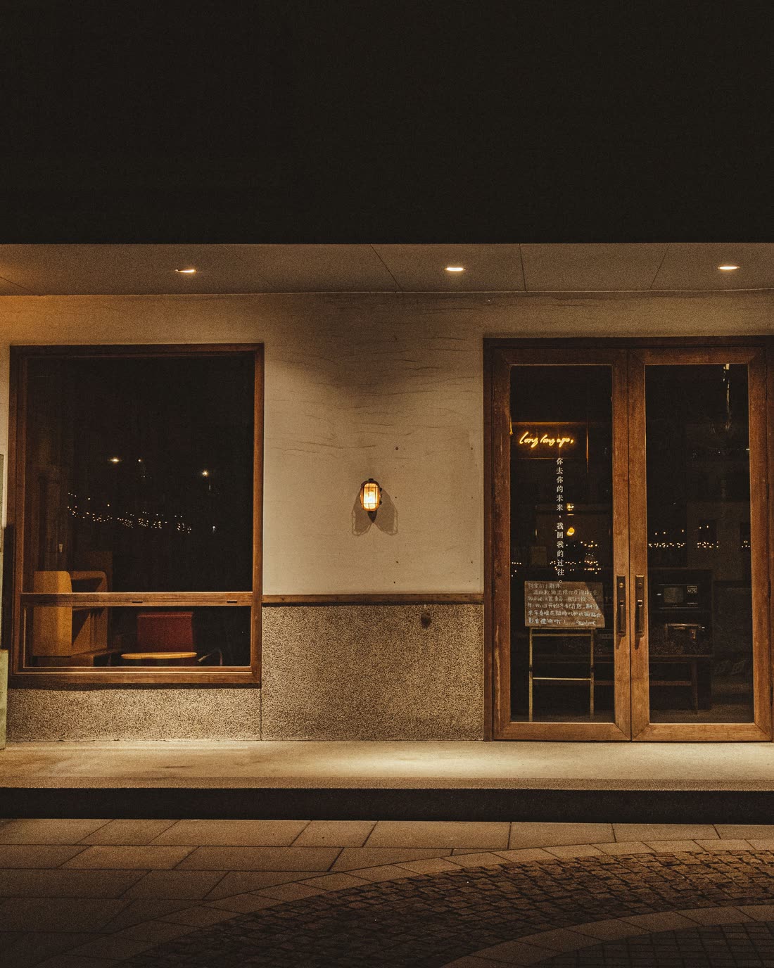 A restaurant frontage at night: two warm windows, wooden doors and wall lamps over wet cobbles.