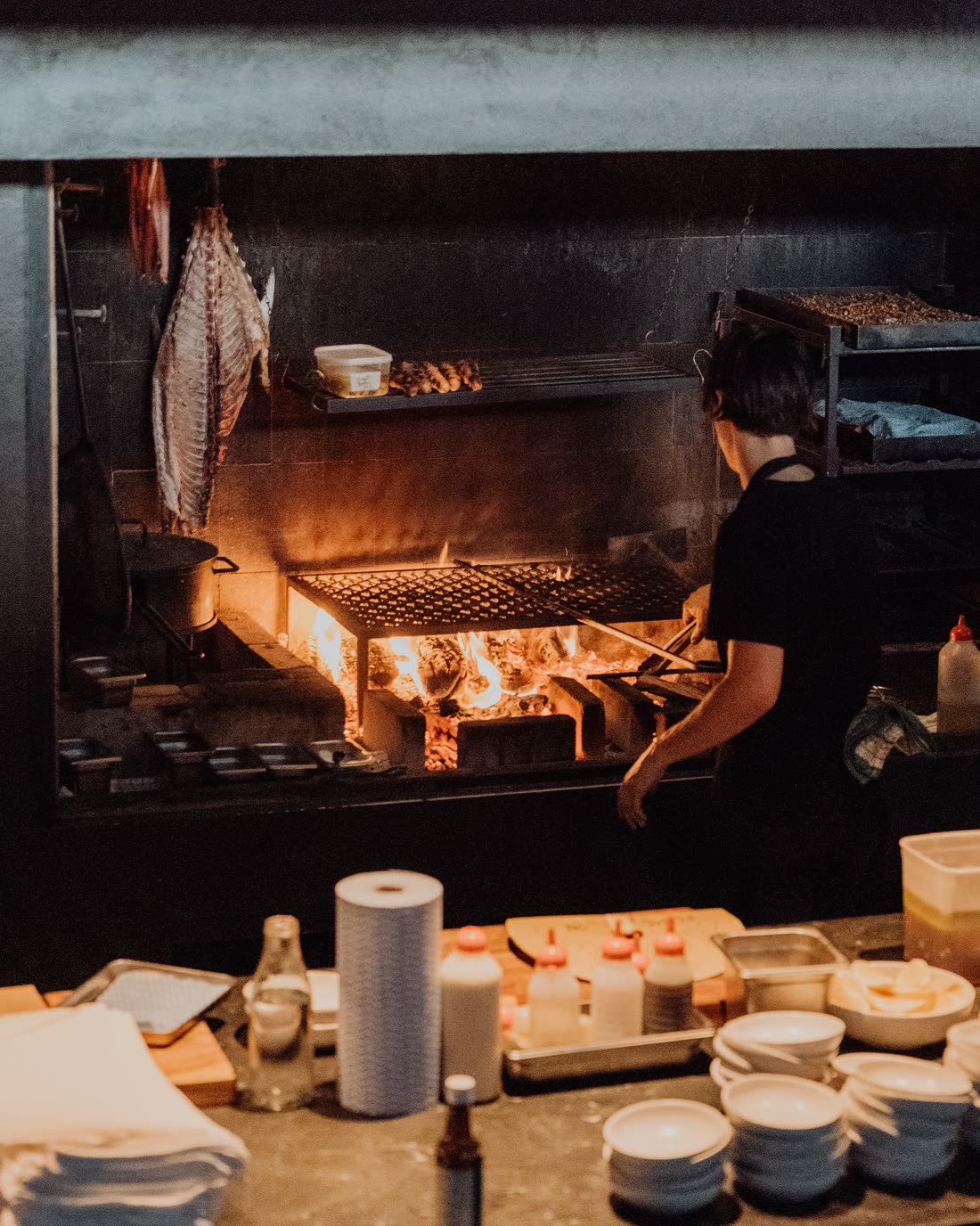 A cook working at a wood-fired grill, lit almost entirely by the flames, with the rest of the kitchen in darkness.